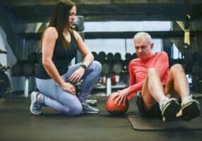 A woman kneels beside an older man seated on a gym floor in Asheville. The man, in a long-sleeve top and shorts, is engaging in core exercises with a medicine ball—an inspiring scene of active senior living. Gym equipment and weights linger in the background.