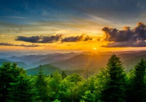 A panoramic view of the rolling Asheville mountains at sunset, with the sun partially hidden behind clouds, casting warm orange and yellow hues across the sky. The foreground features lush green trees under a colorful sky, perfect for anyone considering retirement in scenic serenity.