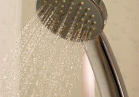 A close-up of a silver showerhead in a Hendersonville senior living facility, with water spraying out in multiple streams against a plain background. The water droplets are visible as they fall.