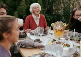 An elderly woman in a red sweater laughs while sitting at an outdoor table, enjoying food and drinks with friends. Surrounded by the lush garden of a Hendersonville senior living community, she savors the joy of retirement and companionship.