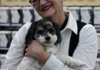 An older woman with short white hair and glasses is smiling while holding a small black and white dog. They are standing in a store in Asheville, with shelves of products and large SAXX signs in the background—a joyful scene that reflects the perks of retirement.