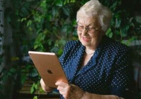 An elderly woman with white hair and glasses is smiling while using a tablet, embodying the joys of senior living. She is wearing a navy blue polka dot blouse, and vibrant green plants form a serene backdrop, reminiscent of retirement days spent in Ashevilles lush beauty.